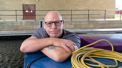 Sofía Luis-Hobbs/BBC Eric Wilson, who is bald and wears black rimmed glasses. He is smiling in the photo while leaning on a narrow boat.