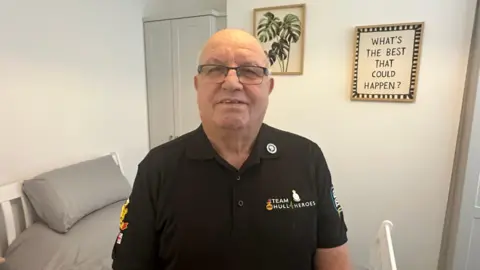 A man stands in a modern-looking bedroom. He has cropped white, thinning hair and wears dark-rimmed glasses and a black polo shirt with the logo "Team Hull 4 Heroes" on it. The bedroom has white walls, a grey wardrobe and a white bed with grey bedding. A picture of a green plant hangs from the wall, while another decoration carries the saying: "What's the best that could happen?"