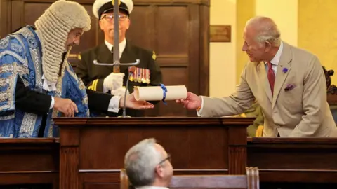 PA Media Skelly, ataviado con la túnica ceremonial del Presidente, entrega el pergamino al Rey, con el portador de la espada detrás y el Presidente de la Cámara observando.