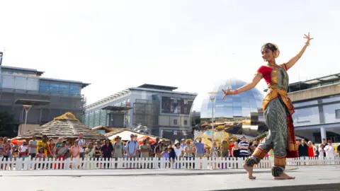 Paul Cox A woman in traditional Indian clothing, which is red, gold and silver, dancing in front of the Planetarium in Millennium Square