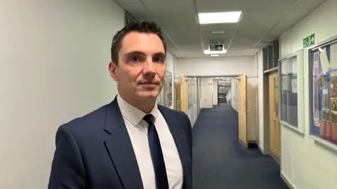 BBC Dominic Wilkes standing in an empty hallway. He is wearing a navy suit jacket with a white shirt and navy tie. The corridor is large behind him and there is a number of signs on the walls.