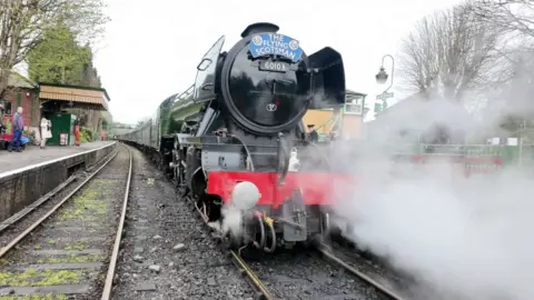 The Flying Scotsman has pulled up at a railway platform. It has a black front with a blue sign reading 'The Flying Scotsman' and steam coming out of the side.
