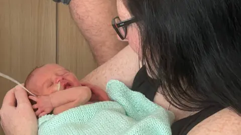 Becky holding baby Noah. Her face is turned away from the camera and she is looking down at him. Becky has dark hair and glasses. Noah is wrapped up in Becky's arms in a mint green blanket. His eyes are closed, his mouth is open and he has one hand up by his ear. He has a feeding tube in his nose. 