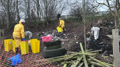 People in protective suits work on a clearup of waste on a woodland verge close to a road. 