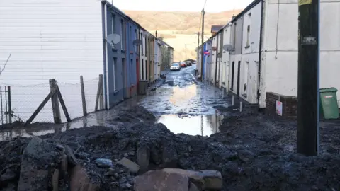 BBC The entrance to a street in daylight is blocked by rubble and mud. Beyond the rubble, water stands in the street and the mountain behind is visible in the background