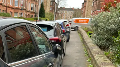 A row of parked cars lines a narrow residential street beside red‑sandstone tenement buildings. A low stone wall runs along the right side with dense shrubs and plants behind it. An orange‑and‑white van is visible farther down the street near more tenement buildings under an overcast sky.