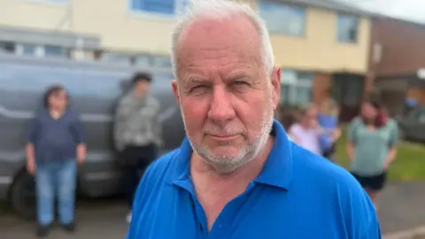 A man with grey hair and a beard and a blue polo neck T-shirt with blurred shapes of people behind him