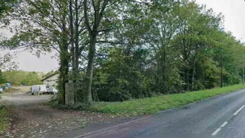 Land behind a row of trees, as seen from a road. There is a single storey building, a white van and a white car seen through a gap in the trees on the land.
