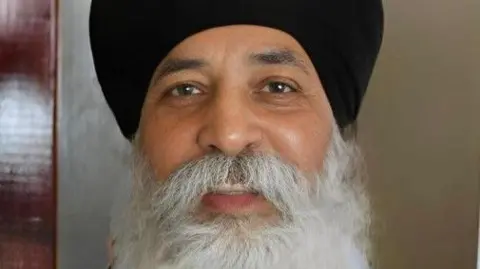 Balhar Singh Gill is wearing a black turban. He has a grey beard and brown eyes.