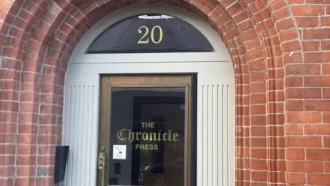 The door to the building, which is glass and says 'The Chronicle Press' in gold letters on it. The brick around the door is red. 