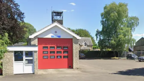 A small building with a large red door and a sign above the door saying Helmsley Fire Station stands in the middle of a nearly empty car park. Trees and a building that looks like a house in the background.