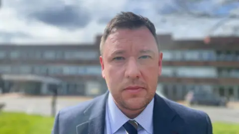 Head and shoulders of Det Insp Alan Sharp who is looking at the camera. He is wearing a blue suit jacket and a blue and white checked shirt with a stripey blue, brown and white tie. He is standing outside a building that is blurred in the background.