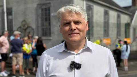Kieron has neat grey hair, roughly parted. Behind him is a chirch and he is wearing a pale shirt.