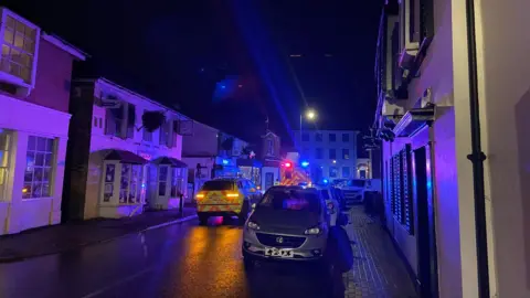 A night time scene showing a village road with two ambulance vehicles parked in the middle of it and a parked car on the side of the road. The ambulance has its red and blue lights lit up but the street is otherwise dark