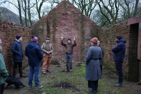 The Hugo Burge Foundation/Stewart Attwood Andrew Goldsworthy stands in derelict farm building that is missing its roof; he is speaking to several people surrounding him 