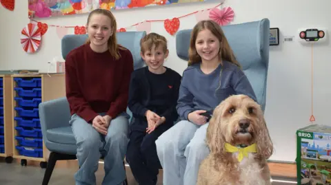 Three children smile at the camera as they sit on a blue chairs in a white-painted room with colourful decorations. In front of them sits a golden coloured dog with floppy ears wearing a yellow bow round its neck.