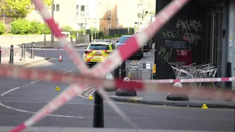 Red police tape crosses the road. Silver chairs are on the pavement opposite, inside the police cordon with a police car with fluorescent yellow marking parked beyond it.