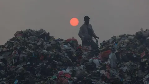 Getty Images A person walks amid piles of rubbish with a low red sun in the sky behind him