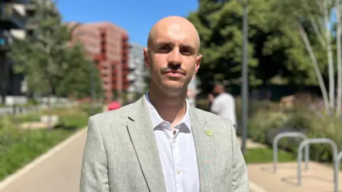 Man with shaven head, light grey suit jacket and white shirt. Stands in a park area with bike rack on his right and greenery on his left.