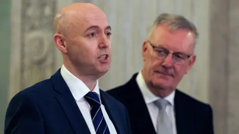 PA Media Burrows, bald and wearing a navy suit, has his lips separated but teeth clenched as he speaks. Behind him, slightly out of focus, Mike Nesbitt, wearing rectangular glasses, looks on, listening.
