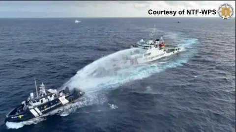 The National Task Force, a Navy Blue Chinese Ship for the West Philippine Sea, is firing a water cannon towards a large white and red Philippine vessel. They sit in the sea, no land is visible. Two other ships can be seen in distance