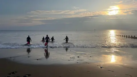 People paddling out to sea on a wide sandy beach with the sunlight low and warm.