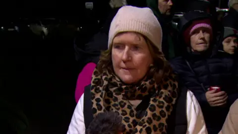 Patricia Dunne is outside with people standing around her. It's nighttime. She's wearing a hat and a cheetah print scarf.