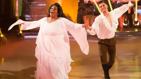 Alison Hammond su una pista da ballo esibendosi con il partner Aljaz Skorjanec. Lei indossa un lungo abito rosa e fluente, lui pantaloni neri e una camicia bianca con volant. Le tiene la mano ed entrambi hanno le braccia spalancate e sorridono ampiamente.