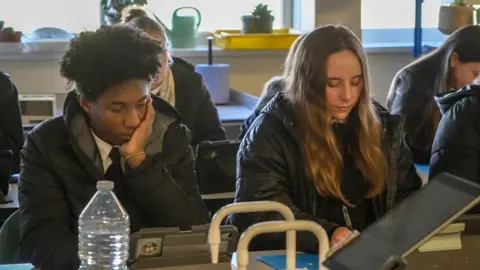 In a classroom, two students sit in their coats while working. One male student with black hair is leaning on his hand while in a puffer jacket. He is sat next to a female student who has long mousey brown hair who is writing with a pen on paper and is wearing a parker style coat. 