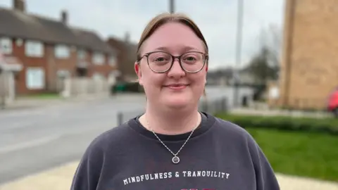 Ben Mellor/BBC Image of Emily. She is wearing glasses and has a blue jumper on. She is stood on her driveway.