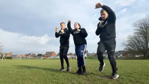 BBC Three people wearing black jumpers taking part in fitness training on grass.