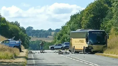 A yellow and brown bus on the side of the road. The left hand side of the bus is damaged, with debris on the road. A white van can be seen on the other side of the road. A person stands in the middle of the road, with two vehicles in the distance. 