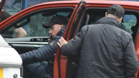 National Crime Agency Image of Arsen Feci in the front passenger seat of a red car. There is a another man with his back to the camera with the back-left passenger door open.