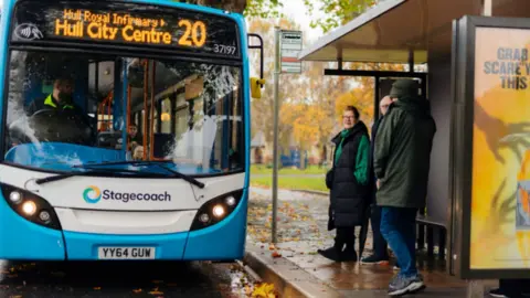 Three people stand at a bus shelter with a blue and white single deck Stagecoach bus number 20 to Hull city centre parked alongside