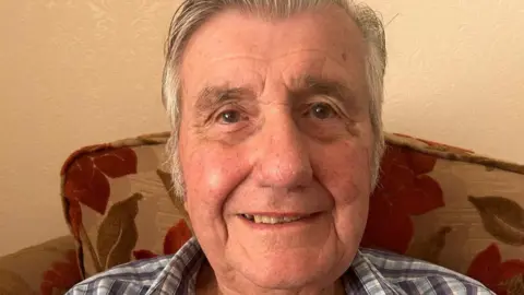 Helen Russell David Judd sits in a living room and smiles at the camera. He has combed grey hair and a check shirt.