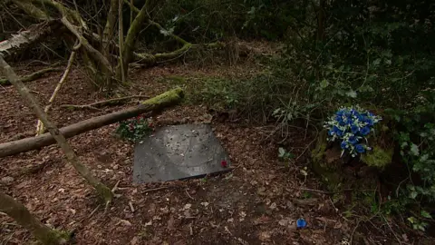 A flat stone marks the grave in the woods, with some blue flowers lying next to it. There is greenery and broken branches all around.