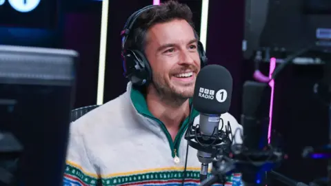 Jonathan Bailey smiling in front of a Radio 1 branded microphone and headphones in the studio.