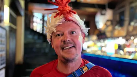 George Carden/BBC A man in a red shirt and a red and white mohawk.