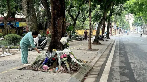 A woman in mint green clothes bends over a ring of flower bouquets arranged around the trunk of a roadside tree