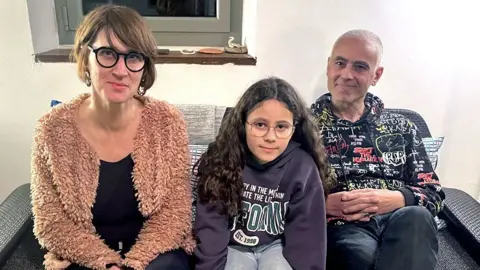 Marion, Mael and Farid sit on a grey sofa. They are women wearing short brown hair and round glasses wearing beige jumpers, young girls with long brown hair, glasses and purple sweater, and older men with white hair and black sweater