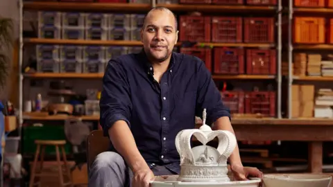 A man wearing a dark blue shirt and grey jeans is sitting in a ceramic studio. There is a clay model of a crown in front of him. 