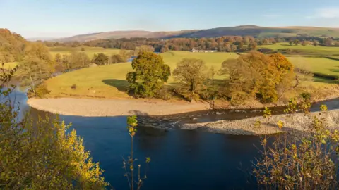 visitlakedistrict.com A river in front of a pebbled river bank and green fields. Hills can be seen in the background alongside trees. The leaves on the trees are just turning orange.