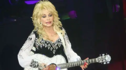 Getty Holding a glittering white guitar, Dolly Parton stands on stage wearing a black dress with wide sleeves.