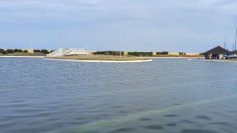 Redcar Model Boat Club The refilled Redcar boating lake. A white footbridge is crossing it at the centre. The boathouse cafe is on the right side of the lake. Two windmills are visible in the distance.