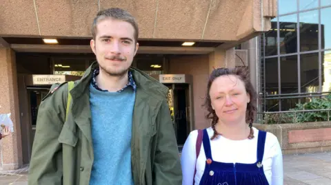 Noah is wearing a green jacket and a blue jumper and has short hair and a beard. Lucy Timlin has brown hair in two plaits and is wearing a white long sleeved t-shirt and blue dungarees. Both are stood outside Liverpool Crown Court and are smiling at the camera.