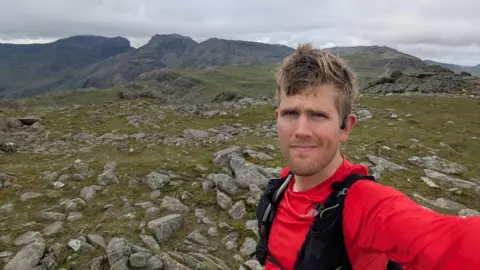 A selfie taken by Alex Staniforth. He has fair hair and stubble and is wearing a red top. He is standing on a Lake District fell with several other peaks visible in the background.