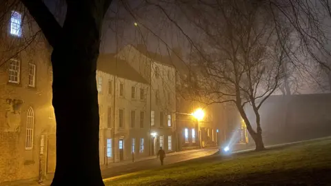 BBC Weather Watchers/DurhamAnne A shadowed figure walking along the a street. To the left is a row of old, white houses with large windows. Some of the lights on the lower floors are on. There are orange lights lighting up the street. There is a patch of grass on the right of the street which is on an incline. There is a shadowed tree trunk in the foreground.