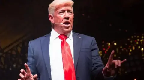 Mike Osman Donald Trump impersonator, Mike Osman, with a blue suit and crimson tie talking to an audience with a head set microphone. 
