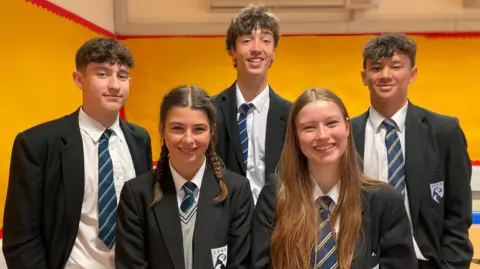 The image shows five students wearing school uniforms standing together indoors. There are bright yellow and red classroom display boards behind them. The students are wearing school uniform including blazers which feature badges on the breast pockets.