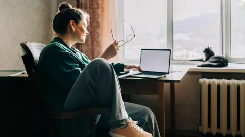 Getty Images Woman holding a pair of glasses sits at a desk in a flat with a small laptop in front of her and a black cat sitting on the window sill.